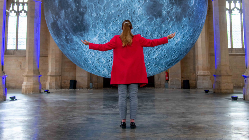 Woman in a red coat standing with her arm stretched in front of the moon sculpture. Woman in a red coat standing with her arm stretched in front of the moon sculpture.