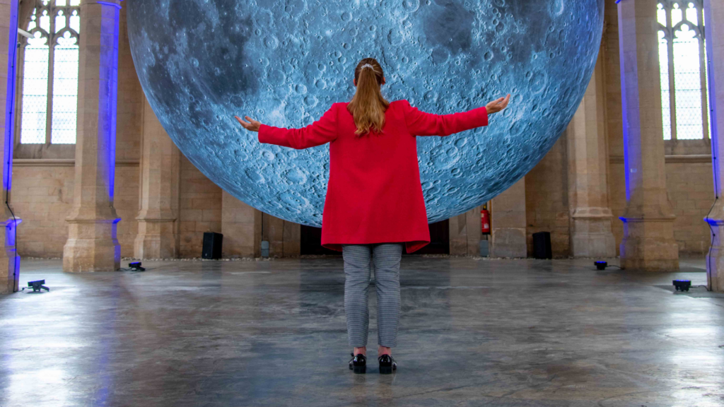 Woman in a red coat standing with her arm stretched in front of the moon sculpture.