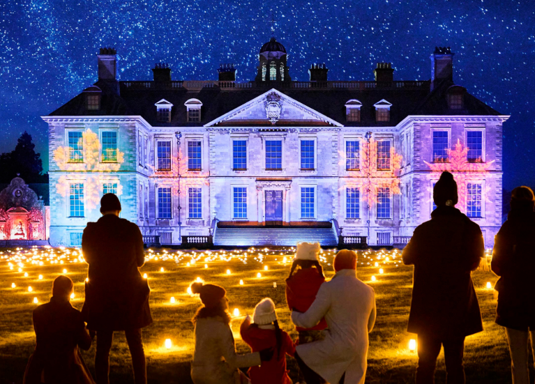 Christmas at Belton - a family looking at Belton House lit up in purple with festive lights on the grounds in front.