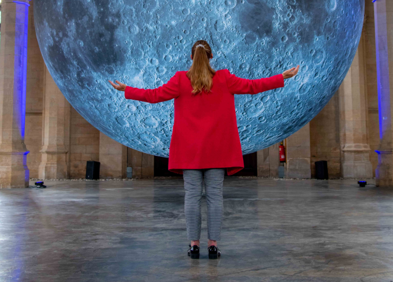 Woman in a red coat standing with her arm stretched in front of the moon sculpture.